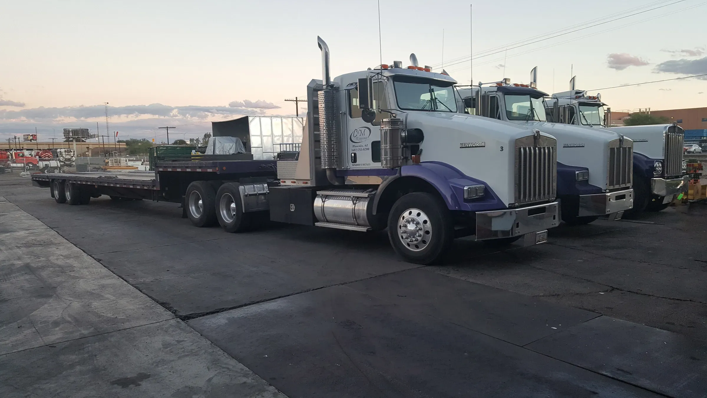 Three C&M Rigging Kenworth tractors with purple fenders and flatbed trailers parked at the Phoenix yard at dusk.